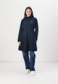Impermeabile blu navy con vita regolabile, chiusura a zip e cappuccio. Realizzato in materiale resistente all'acqua. Abbinato a jeans a gamba larga di un delicato blu.