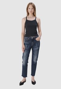 Débardeur noir à côtes associé à un jean bleu foncé légèrement déchiré avec des ourlets effilochés. Modèle portant des ballerines noires, posant devant un fond uni.