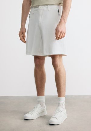 Mann trägt weiße knielange Shorts mit seitlichen Reißverschlusstaschen, weiße Socken und weiße Sneakers auf grau meliertem Boden.