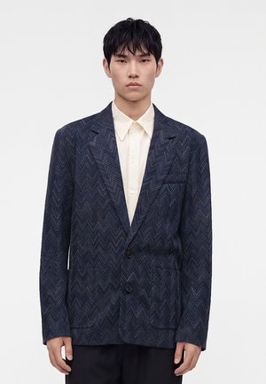 Jeune homme aux cheveux noirs courts portant un blazer bleu foncé texturé par-dessus une chemise blanche boutonnée sur un fond blanc uni.