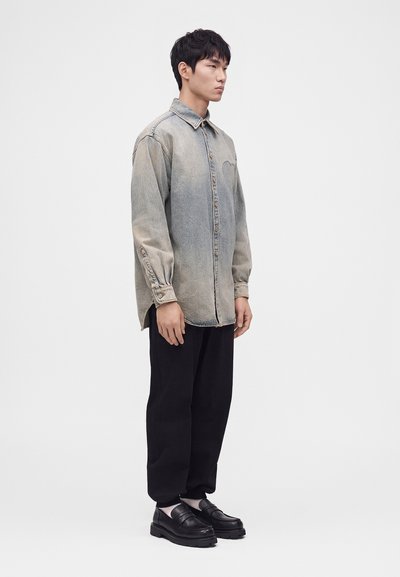Homme debout portant une chemise en jean délavée à boutons, un pantalon noir fuselé, des chaussettes blanches et des mocassins en cuir noirs sur un fond clair uni.