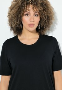Top negro de manga corta hecho de una tela suave, con un escote redondo y un corte relajado. Modelo con cabello rizado y pendientes llamativos.