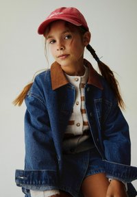 Džinsinė striukė su ruda korduroko apykakle, kreminė dryžuota megztinis ir raudona kepurė su užrašu „gelato“. Plaukai pinti kasomis, neutrali fonas.