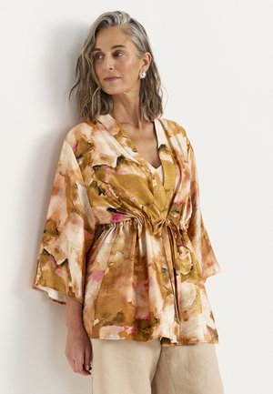 Kobieta o szarych, sięgających ramion włosach nosi luźną, kwiecistą, w ziemistych tonach, kimono zawiązane w pasie oraz beżowe spodnie.