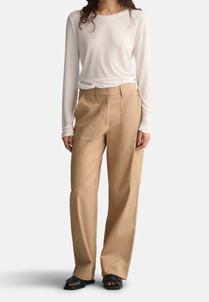 Kobieta w białej koszuli z długim rękawem, luźnych beżowych spodniach z dużymi kieszeniami i czarnych sandałach z odkrytymi palcami, stojąca na jednolitym tle.