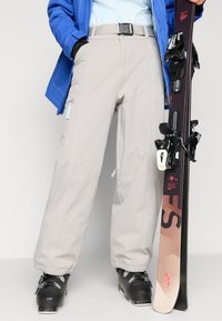 Calças de esqui cinza claro com um corte relaxado, apresentando costura em contraste, bolsos laterais e um cinto, combinadas com botas de esqui pretas e uma prancha de snowboard.