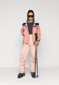 Conjunto de ski com uma jaqueta impermeável rosa e calças rosa claro. Botas de ski pretas e bastões complementam o visual, com um forro interior cinza.
