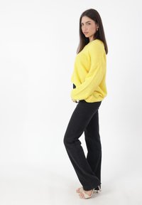 Gelb gestrickter Pullover in lockerer Passform, schwarze hochtaillierte Jeans und beige Absatzsandalen. Die Textur des Pullovers wirkt weich.