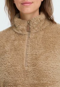 Beiger Fleece-Pullover mit hohem Kragen, halbem Reißverschluss und plüschiger Textur. Verfügt über ein kleines Leder-Branding-Tag in der Nähe des Reißverschlusses.