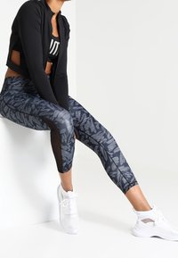 Sort cropped jakke over en sort og grå mønstret sportstop. Grå leggings med abstrakte mørkegrå mønstre og mesh-indlæg. Hvide sneakers.