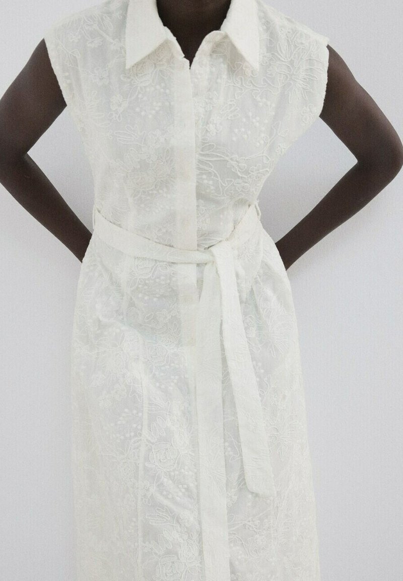 Robe blanche sans manches avec broderie florale, col et ceinture à la taille, portée par une personne les mains sur les hanches sur un fond uni.