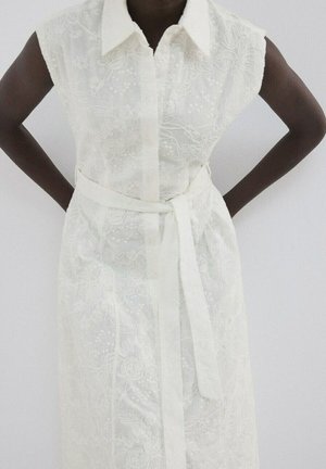 Vestito bianco senza maniche con ricamo floreale, colletto e cintura in vita, indossato da una persona con le mani sui fianchi su uno sfondo uniforme.