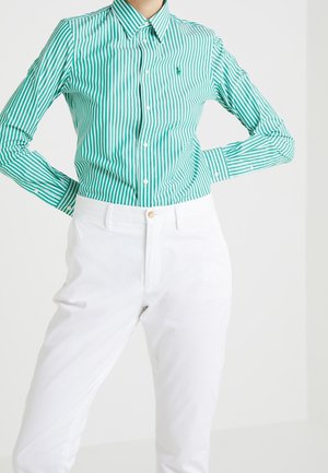 Osoba ubrana w koszulę na guziki w zielono-białe pionowe paski, włożoną do białych spodni z wysokim stanem na jednolitym tle.