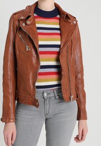 Person som bär en brun läderjacka över en ribbad, flerfärgad randig tröja och ljusgrå jeans, stående mot en neutral bakgrund.