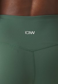 Leggings sportivi verdi realizzati in un materiale liscio ed elastico. Logo bianco "ICIW" prominente sulla vita e dettagli delle cuciture discreti.