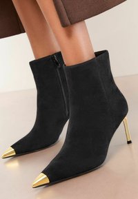 Fekete velúr bokacsizmák hegyes orral és fényes arany részletekkel az orrán. Oldalra nyíló cipzárral és vékony, magas sarokkal rendelkezik.