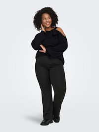 Pull en tricot noir avec découpes aux épaules, grand nœud décoratif, et coupe ample, associé à un pantalon noir droit et des chaussures noires.