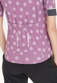 Lila Kurzarmoberteil mit weißem kreisförmigem Muster, elastischem Taillenbund und reflektierenden Streifen. Kombiniert mit schwarzen Hosen.