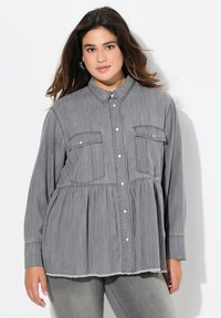 Camicia grigia con bottoni e design a peplo, dotata di due tasche sul petto, maniche lunghe e orlo sfilacciato. Realizzata in un tessuto morbido e texturizzato.