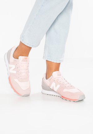 Personne portant des baskets New Balance rose clair et gris, associées à un jean cropped bleu clair sur un fond blanc.
