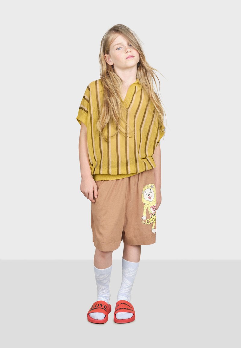Gulstribet kortærmet top, brune shorts med løvegrafik. Hvide knæhøje sokker, røde sandaler med sort tekst. Enkelt, afslappet design.