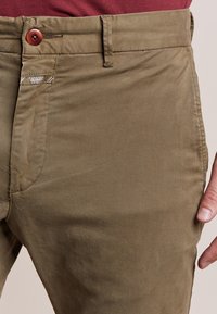 Pantalons khaki en tissu de coton lisse, avec fermeture à bouton et détails de poches latérales. La couleur est un vert-brun atténué, offrant un look décontracté.