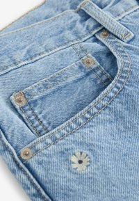 Jeans de mezclilla azul claro con un pequeño detalle de flor bordada en el bolsillo trasero, con remaches de metal plateado y costuras en contraste.