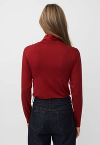 Rode top met lange mouwen en een losse pasvorm, voorzien van een hoge halslijn en zachte textuur. Combinatie met donkere spijkerbroek met achterzakken.
