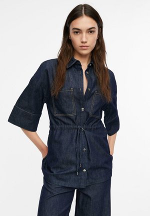 Junge Frau trägt ein dunkelblaues Jeanshemd mit Knöpfen und vorderen Taschen sowie passende weite Hosen und steht mit den Händen in den Taschen.