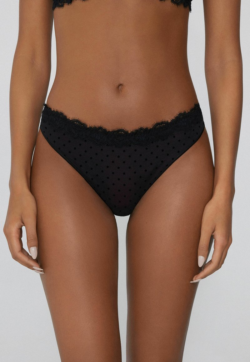 String noir à pois blancs avec une taille en dentelle festonnée. Tissu doux et transparent avec une texture lisse et un design à couverture minimale.
