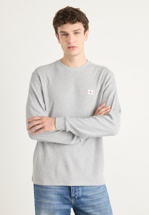 Grå langermet t-skjorte laget av teksturert stoff med rund hals; har en liten logo-patch på venstre bryst. Brukt med blå jeans.