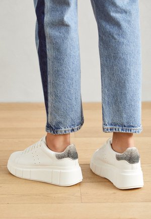 Person iført lyseblå jeans og hvide platform-sneakers med metallic sølv hældetalje, der står på trægulv.