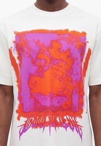 Λευκό T-shirt με μεγάλο κόκκινο και μοβ αφηρημένο σχέδιο με μάτια και μία νυχτερίδα, με στιλιζαρισμένο κείμενο στο κάτω μέρος.