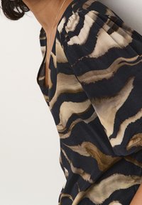 Haut à manches courtes avec un motif ondulé noir, beige et marron et un décolleté en V profond. Le tissu semble léger et légèrement texturé.