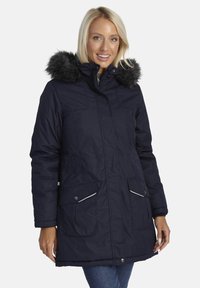 Smaidoša sieviete, kas valkā tumši zilu ziemas parku ar melnu mēteļa apkakli un priekšējiem kabatām, stāv uz baltas fona.