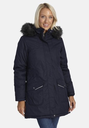 Lachende vrouw in een marineblauwe winterparka met een capuchon van zwart bont en voorflapzakken, staand tegen een witte achtergrond.