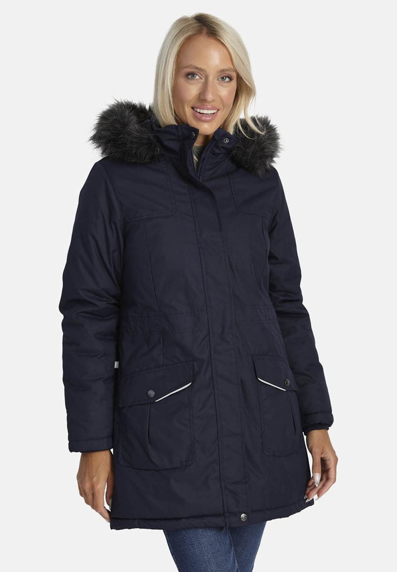 Smaidoša sieviete, kas valkā tumši zilu ziemas parku ar melnu mēteļa apkakli un priekšējiem kabatām, stāv uz baltas fona.