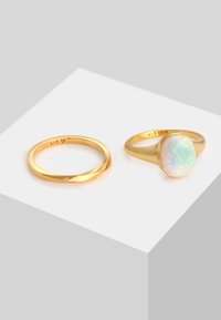 Guld-färgade ringar visade på en ljusgrå yta; en slät ring och en som har en oval opalsten med flerfärgade prickar.