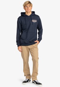 Sudadera azul marino con un bolsillo tipo canguro en el frente y capucha, que presenta un logotipo rectangular de Billabong. Combinada con pantalones caqui y zapatillas negras.