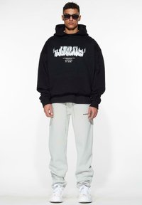 Zwarte oversized hoodie met witte grafische tekst, gecombineerd met lichtgrijze joggingsbroek. Het model draagt ook donkere zonnebril en witte sneakers.