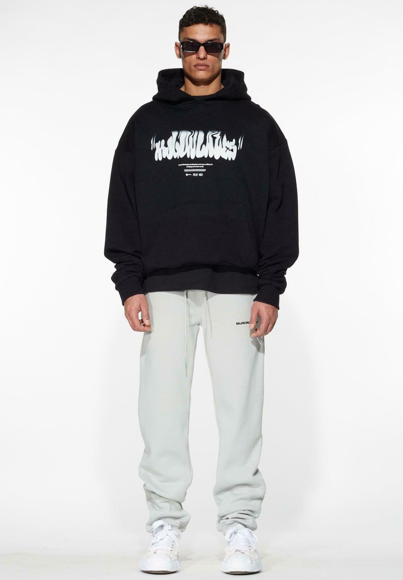 Zwarte oversized hoodie met witte grafische tekst, gecombineerd met lichtgrijze joggingsbroek. Het model draagt ook donkere zonnebril en witte sneakers.