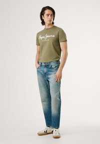 Camiseta de algodón verde oliva con texto blanco "Pepe Jeans London", combinada con jeans de pierna recta azul claro y zapatillas blancas con detalles negros.