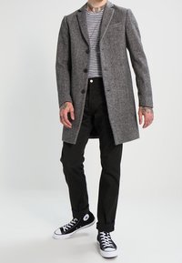 Cappotto di lana grigio con rever, bottoni neri, indossato sopra una camicia a righe bianco e nera e pantaloni neri, con sneaker nere.