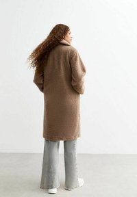 Brun, fluffig teddyjacka med hög krage, knälång design och vida ärmar, i kombination med ljusgrå utsvängda jeans och vita skor.