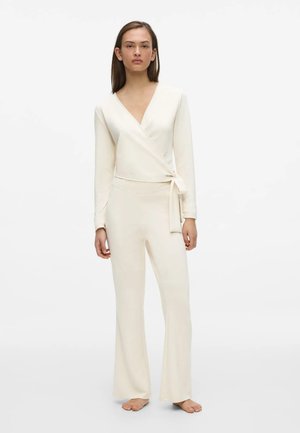 Femme debout pieds nus portant un haut crème à manches longues de style cache-cœur avec un nœud sur le côté et un pantalon large assorti sur fond blanc.
