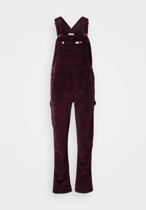 Pantalones de tirantes burdeos de pana con correas ajustables, bolsillos frontales y un corte relajado. Textura acanalada distintiva y acentos de hardware de latón.