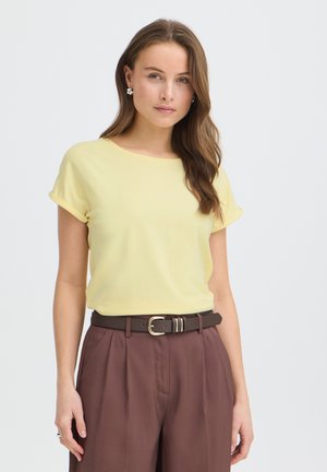 Hosszú barna hajú nő, aki sárga rövid ujjú felsőt, barna magas derekú nadrágot és sötét övet arany csattal visel, fehér háttér előtt áll.