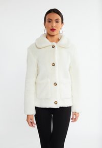 Hvid faux fur jakke med krave, fem sorte knapper og to forlommer. Slim fit med en blød tekstur, båret over sorte bukser.