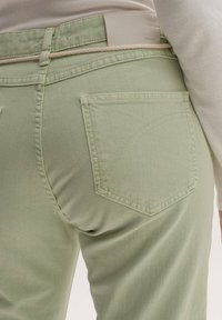 Person i ljusgröna jeans med sömmar på bakfickorna, en beige flätad bälte och en ljusbeige topp instoppad.