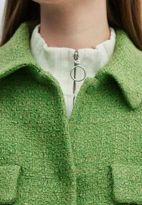Green textured jacket with a collared neckline, featuring front pockets, a silver zipper pull, and layered over a white ribbed top.